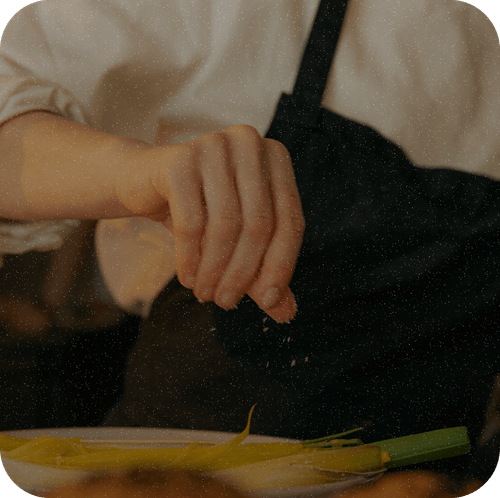 Photographie : un cuisinier assaisonne une assiette en plein service, les deux mains prises.